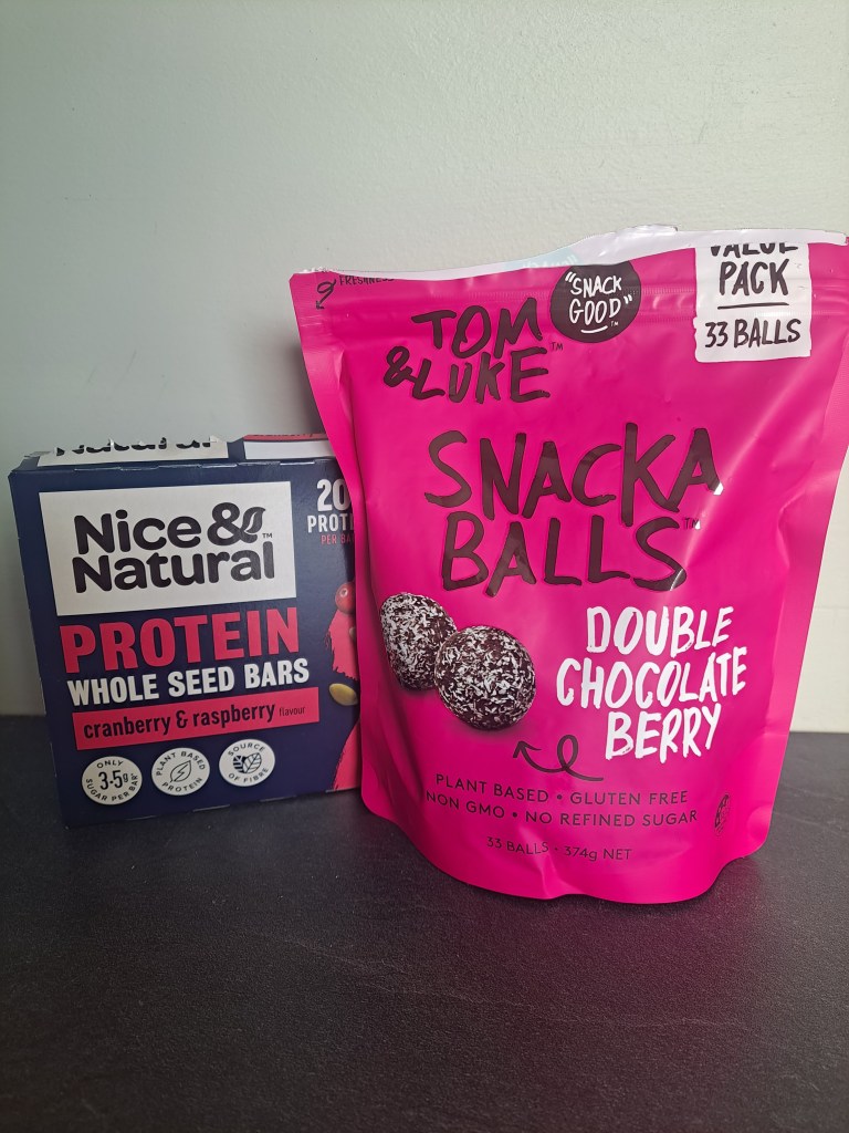 Image of two snack items: a box of Nice & Natural protein whole seed bars with cranberry and raspberry flavor, and a bag of Tom & Luke Snacka Balls in double chocolate berry flavor, placed on a flat surface.