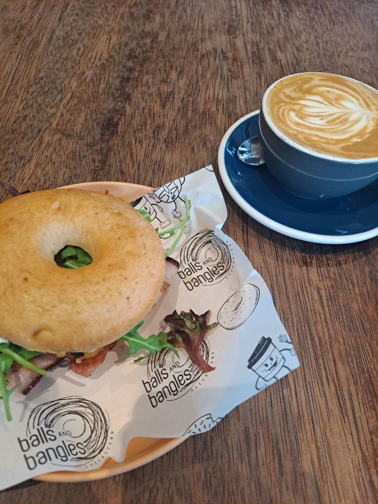 A gluten-free bagel sandwich with greens and bacon, accompanied by a latte on a wooden table at Balls and Bangles café in Queenstown.