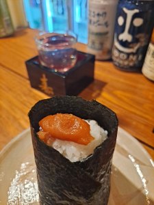 A close-up of a sushi roll wrapped in seaweed, filled with rice and topped with orange fish roe. In the background, a small glass of sake and various beverage cans are visible on a wooden table.